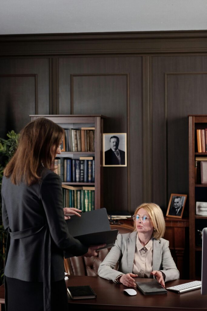 Two businesswomen at a meeting in a book-filled office. Professional and focused setting.